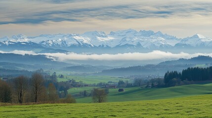 Obraz premium Scenic view of snow capped mountains with mist and green hills under an expansive sky