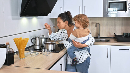 A mother holds her child while tasting food in a modern kitchen, showing multitasking and parenting...