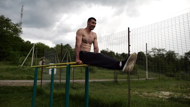 Strong man trains abdominal muscles raising legs on parallel bars at sports ground. Young shirtless sportsman exercising at nature. Sporty athlete working out at nature. Concept of sportive lifestyle