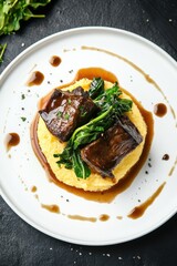 Braised short ribs served over creamy polenta with sautéed greens and savory sauce on a white plate