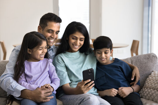 Happy Indian family spend time watching videos or online educational content relaxing on sofa, looking at smartphone screen with joyful expression, enjoy remote talk via videocall app with relatives