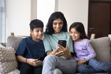 Attractive Indian woman and two little kids using smartphone at home, family spend free time on...