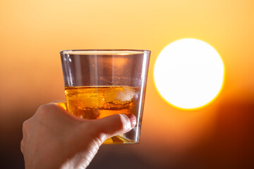 A hand holding a glass of whiskey on the rocks against a golden sunset. Refreshing drink, summer vibes, and relaxation concept. Perfect for lifestyle, travel, and beverage themes