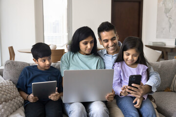 Indian couple and their little son and daughter using diverse modern wireless gadgets seated on couch. Man holds smartphone enjoy new mobile application or game with family, spend leisure on internet