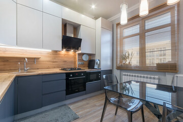 The interior of a modern kitchen. Glass table with chairs made of transparent plastic. Kitchen set in white and gray tones with a wooden countertop.