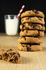 A glass of milk with white and red straw and a stack of chocolate chip cookies. The cookies are piled on top of each other and one is missing a piece