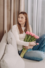 Young woman holding a bouquet of pink tulips, sitting on a cozy couch. Peaceful and thoughtful expression. Soft natural light, modern home interior, spring vibes. 8 March.