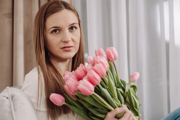 Young woman holding a bouquet of pink tulips, sitting on a cozy couch. Peaceful and thoughtful expression. Soft natural light, modern home interior, spring vibes. 8 March.