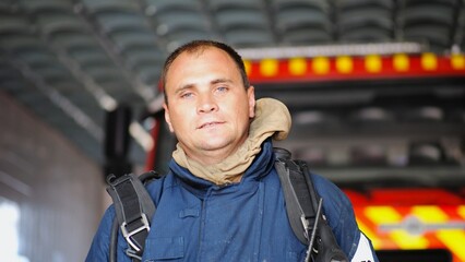Portrait of tired male firefighter standing near a fire engine at department. Young exhausted fireman in protective uniform looking at camera after fire against the background of big red truck