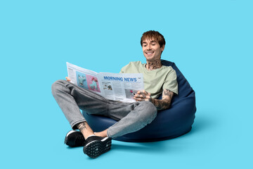 Handsome young man in beanbag chair with newspaper on blue background
