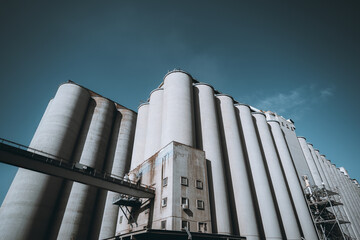 A large industrial grain silo facility with multiple tall, cylindrical concrete storage units under a clear blue sky. The structure includes metal scaffolding, pipelines, and a walkway