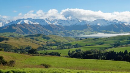 Fototapeta premium Snow capped mountains loom over rolling green hills with mist drifting over peaks under a vast sky filled with clouds