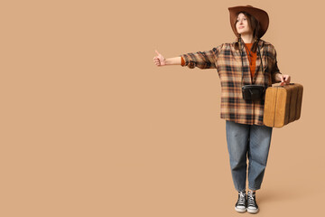 Smiling young woman in cowboy hat with suitcase trying stop car on beige background. Travel concept