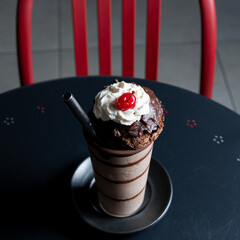 Chocolate milkshake topped with whipped cream, a cherry, and a chocolate brownie, served on a table in a cafeteria