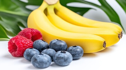 Fresh bananas, raspberries, and blueberries on a white surface with green leaves behind for healthy eating