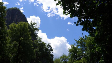 Obraz premium Majestic Mountain Peak and Lush Green Forest Under a Blue Sky with Fluffy Clouds