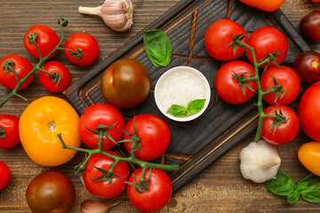 Board with different fresh tomatoes, basil, garlic and bowl of salt on wooden background
