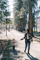 Fototapeta premium Little girl rides a scooter next to her walking mother on the road in a sunny spring forest