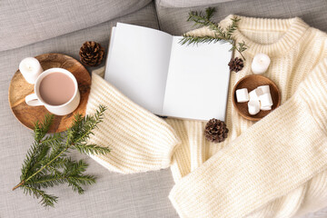 Blank book with cocoa, marshmallows and warm sweater on sofa, top view