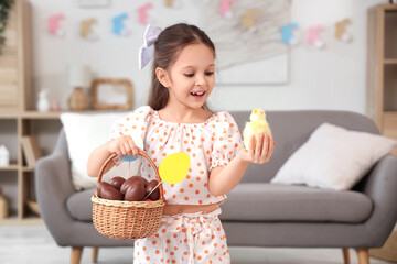 Cute little girl holding wicker basket with sweet chocolate eggs and chick in living room. Easter celebration