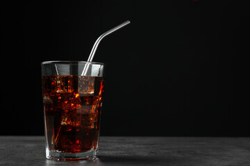 Cold cola with ice cubes and drinking straw in glass on grey table against black background. Space for text