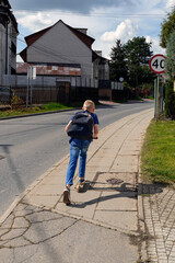 Fototapeta premium Energetic boy riding a scooter along a sidewalk, heading home with a backpack on a sunny afternoon.