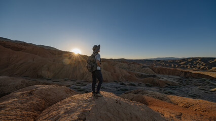 A female tourist in a hat and with a backpack on her back stands on the Martian soil during the golden hour in Mongolia.
