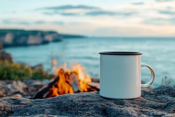 Obraz premium White enamel mug by campfire, ocean view. Perfect for travel, camping, or cozy vibes.