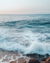 Fototapeta premium Serene ocean vista with gentle waves lapping on sandy beach rocks under a soft pastel sky horizon