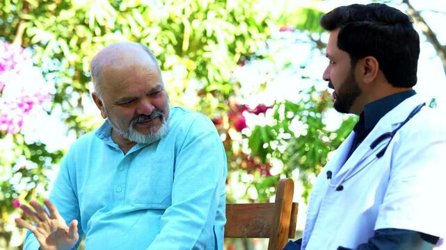 Indian Asian senior male patient in traditional kurta sitting with a young, handsome male doctor in a hospital park as they engage in a lighthearted discussion, sharing smiles and enjoying a friendly 
