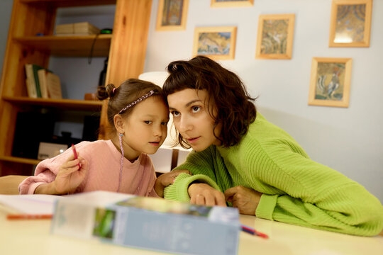 Indoor image of mother and daughter at table, little girl whispering in her ears, asking mom for advice how to draw picture, holding red crayon in hand. Artwork activity and motherhood