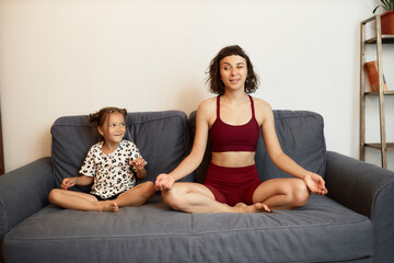 Woman in fitness clothes trying to meditate on sofa while her naughty funny little daughter playing...