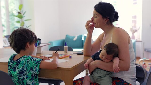 Mother holds her baby while supervising her older child's homeschooling activity at the table, showcasing the family's commitment to education in a calm, home setting