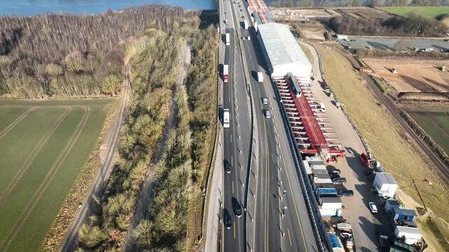 Highway bridge under construction spans canal with multiple lanes of traffic, including trucks and cars navigating temporary road setup marked by traffic cones.