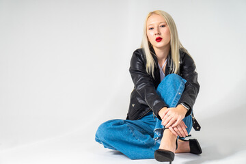 Fototapeta premium Pretty blonde girl sitting on white background. Model is wearing black leaher jacket and blue jeans. Copy space
