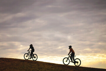 Naklejka premium Two people in helmets riding bicycles up hill at sunset