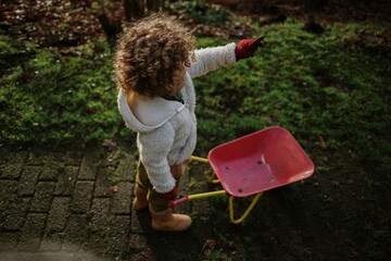 A curly-haired child with a red wheelbarrow exploring the outdoors.