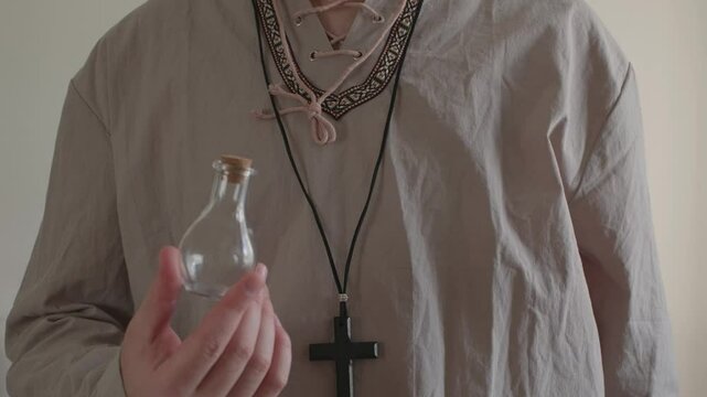 A man with a cross around his neck holding a potion in a red glass bottle	