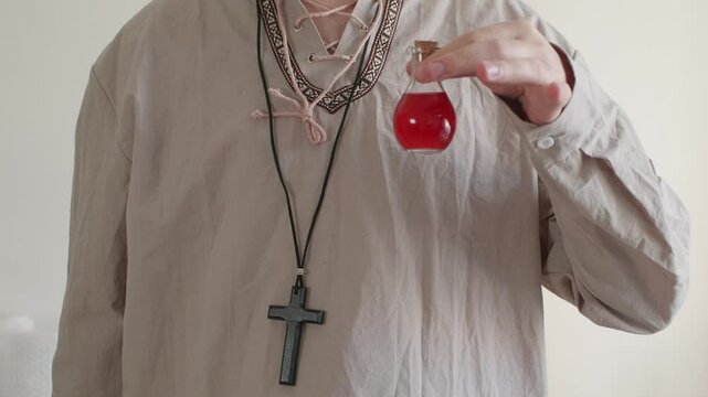 A man with a cross around his neck holding a potion in a red glass bottle	