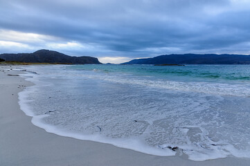 Soft waves create foamy patterns on the sandy beach as the sun sets behind distant mountains, casting a tranquil atmosphere.