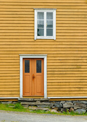 A yellow wooden house features an orange double door and a window above, set against a stone foundation in a rural landscape.