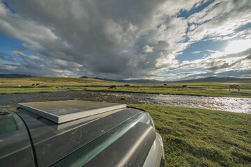 An electric SUV with a solar battery on the hood stands against the background of a green meadow and a river. Solar panel on the background of nature. Alternative green energy.