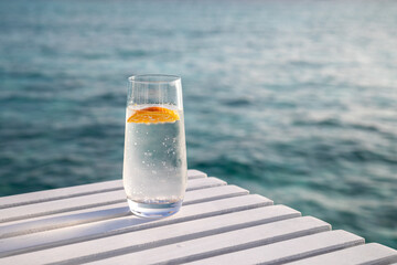 Glass of chilled water with citrus, left of frame, on a white table with sea behind