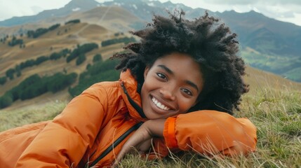 A smiling woman reclines on grass in a mountainous landscape, wearing an orange jacket and black pants, with a backpack nearby.
