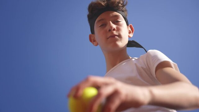 Suggestive low-angle shot of a cute teenage boy tennis player bouncing the ball and serving on a clay court. The blue sky is above him.