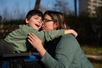 Mother kissing and hugging her son with disability sitting in a wheelchair in a park - cerebral palsy concept