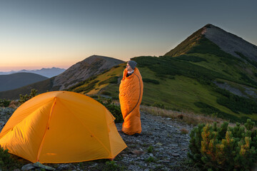 Man standing in a sleeping bag near orange tent at mountain sunrise, enjoying morning view © XArt