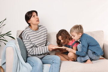 Overwhelmed mother and her naughty children with book on sofa at home