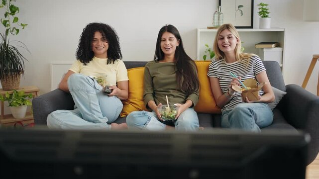 Diverse happy flatmate girl friends watching TV at home while eating healthy meal and takeout food