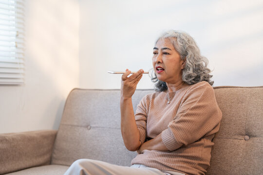 Positive mature woman hold cellphone sits on sofa, record message, talking on speakerphone on smartphone, happy senior use mobile application, enjoy virtual assistant, communicates spend time indoor.